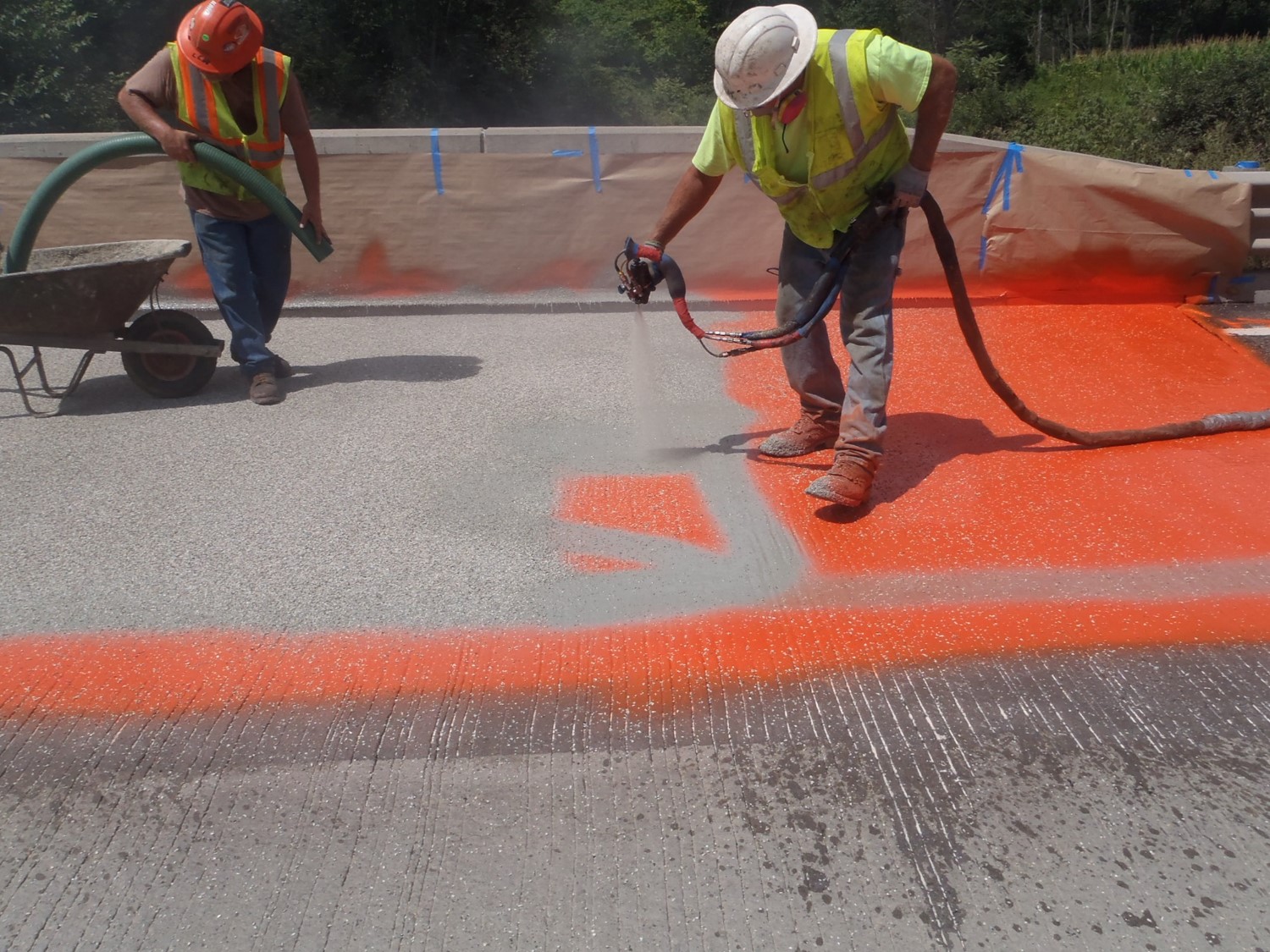 An image of two construction workers in hard hats and yellow safety vests using a sprayer to apply an orange waterproof coating to a bridge deck.